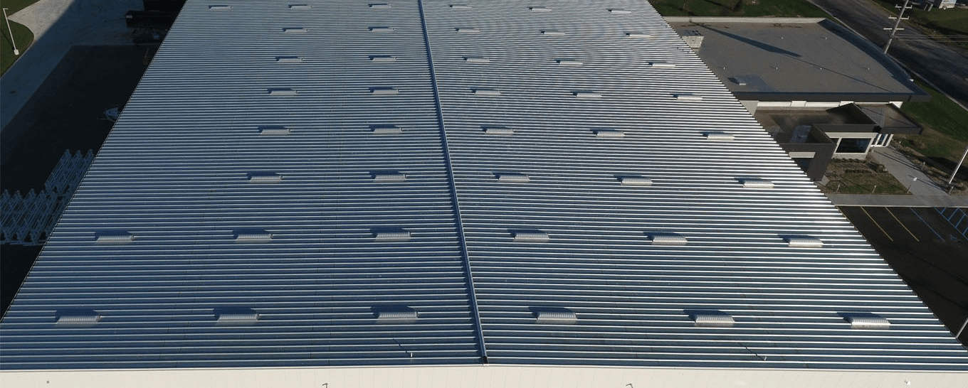 Aerial view of a large metal roof with standing seam panels and evenly spaced skylights running in rows along both sides of the central ridge.